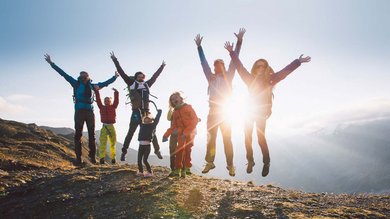 Un gruppo di amici salta in aria gioiosamente sulla cima di una montagna, incorniciati dalla calda luce del tramonto contro il cielo azzurro radioso.