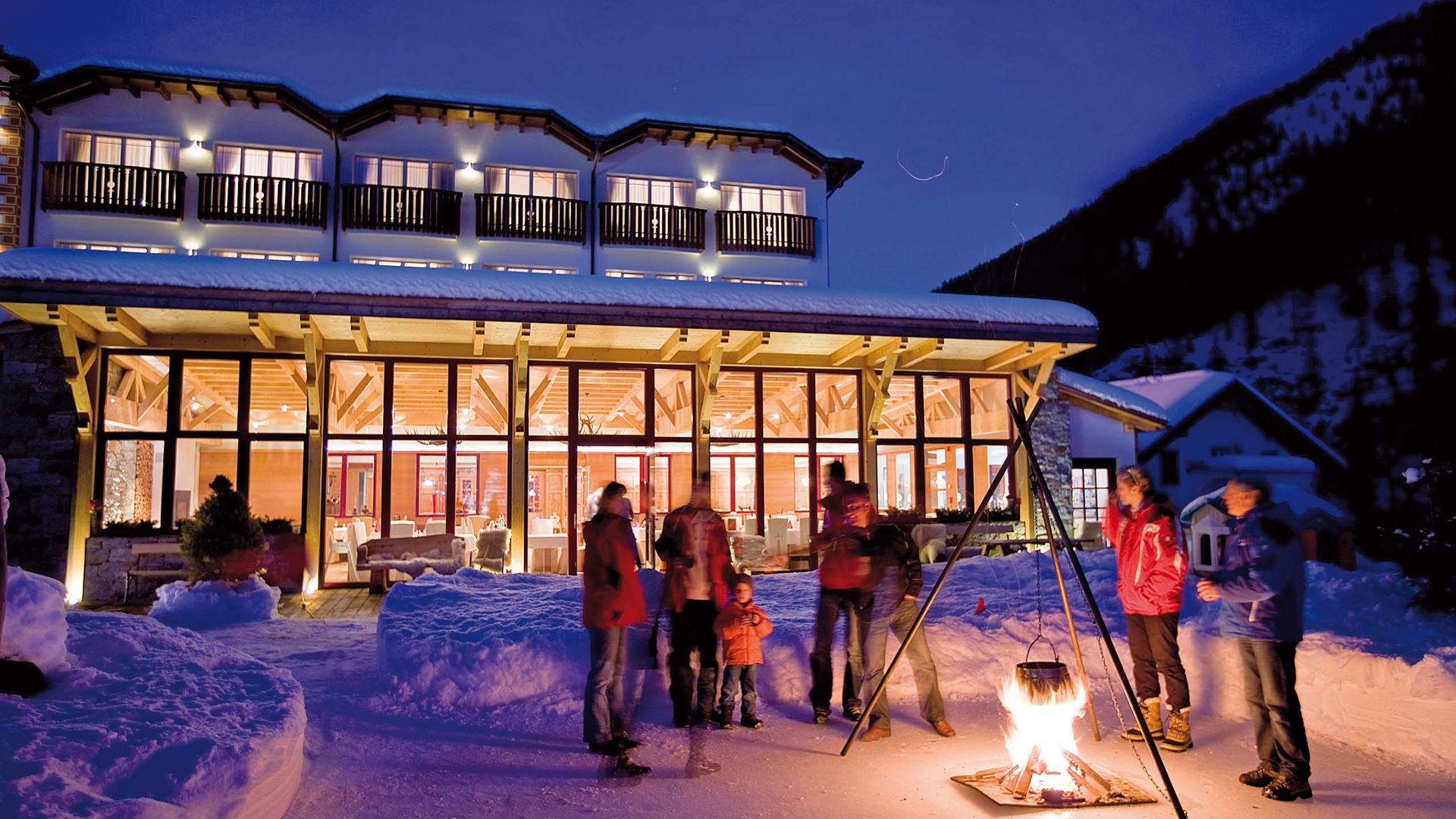 Fröhliche Gäste genießen wärmenden Glühwein im winterlichen Ambiente an der Feuerstelle des Hotel Bella Vista, Südtirol.
